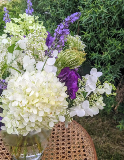 A flower arrangement with purple and white blooms in a glass vase sitting on a rattan table outside near a shrub.