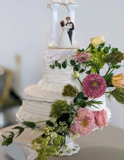 A white, two-tiered wedding cake with a bride and groom statue on top and trailing flowers on the side.