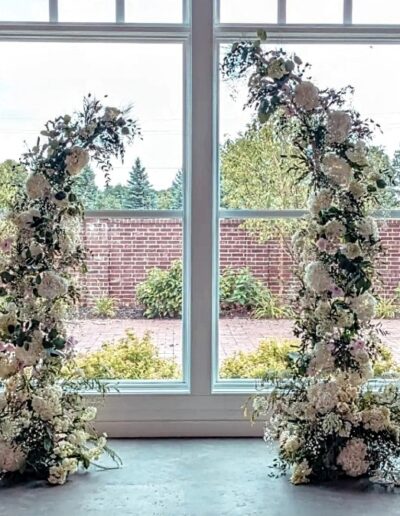 A partial wedding arch made of white flowers in front of a wall of windows.