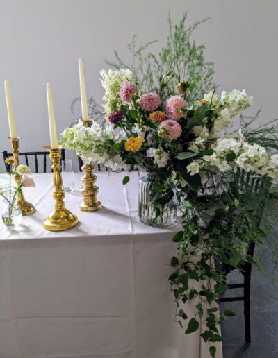 A whimsical flower arrangement on a table next to three golden candlesticks with white tapered candles in them.